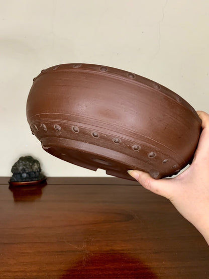 A hand holds a brown, unglazed ceramic pot with raised circular ridges around its rim.