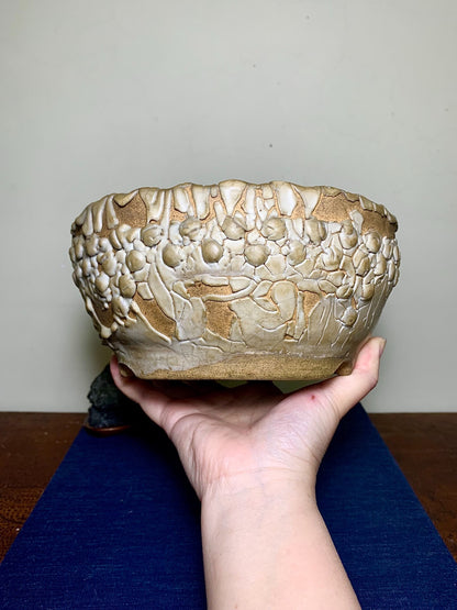 A hand holds a glazed ceramic pot with raised, textured floral patterns in beige and brown.
