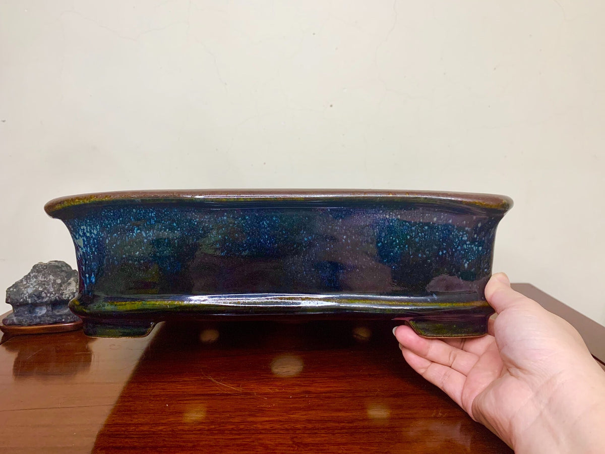 A hand holds a glossy, elongated blue-green glazed pot with speckled glaze and brown rim.