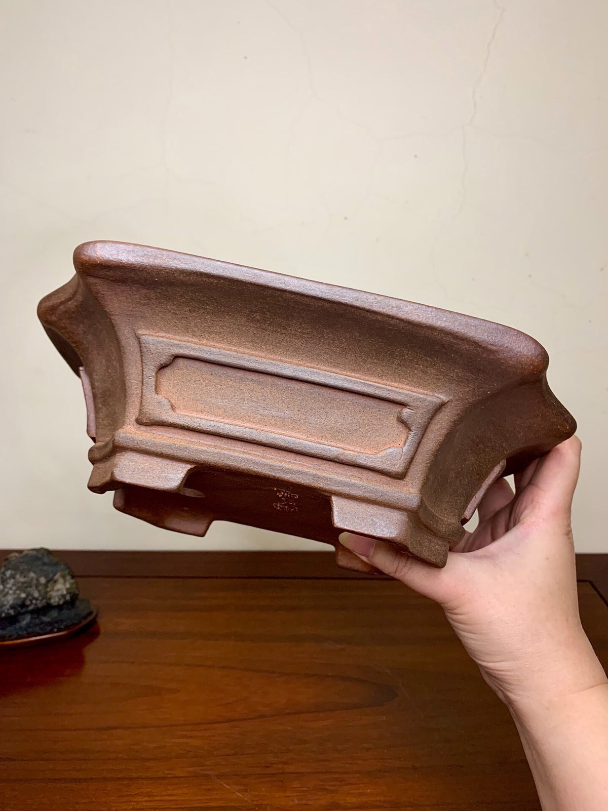 A hand holds a brown, rectangular ceramic pot with carved decorative edges and a matte finish.