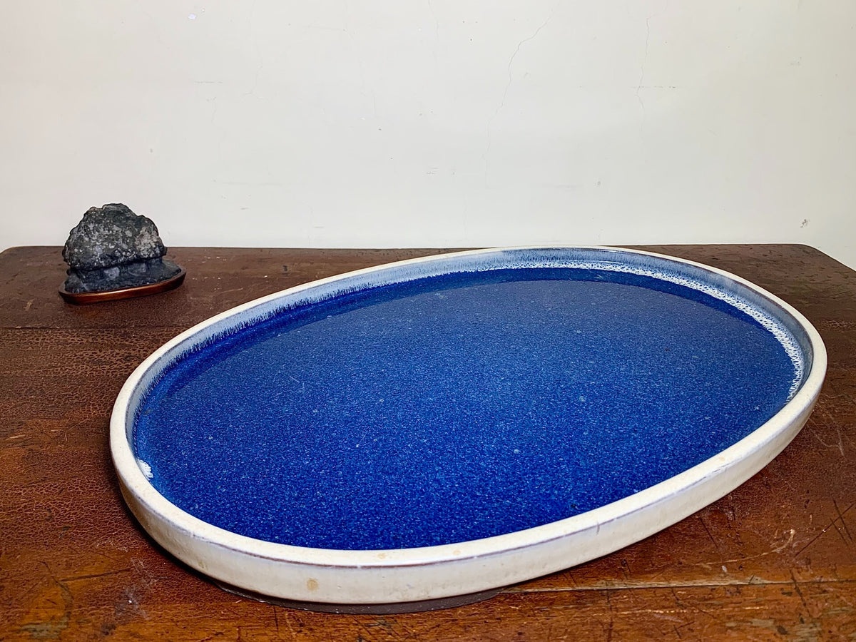 A glossy, deep blue oval tray with a white rim, beside a small dark rock on a wooden surface.