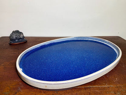 A glossy, deep blue oval tray with a white rim, beside a small dark rock on a wooden surface.