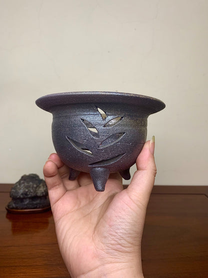 Dark brown glazed pot with leaf-shaped cutouts, held in hand.