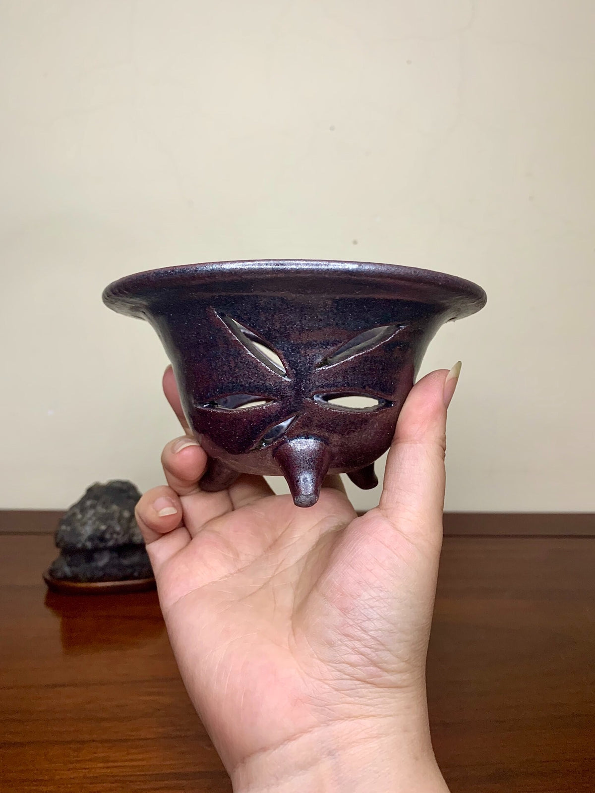 A hand holds a small, dark purple glazed pot with three cut-out eyes and a nose.