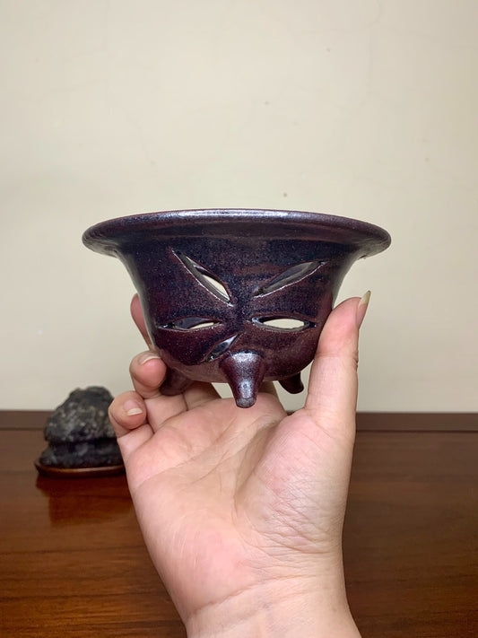 A hand holds a small, dark purple glazed pot with three cut-out eyes and a nose.
