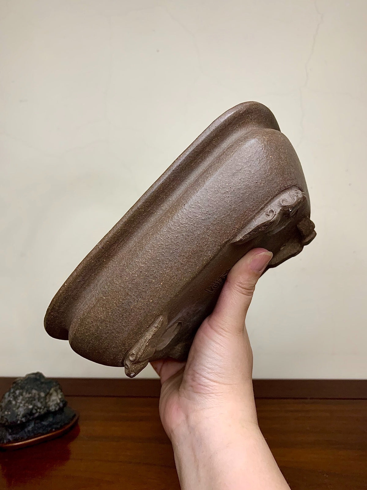 A hand holds a brown, matte-finished, rectangular bonsai pot with three carved feet.