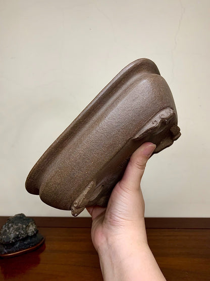 A hand holds a brown, matte-finished, rectangular bonsai pot with three carved feet.