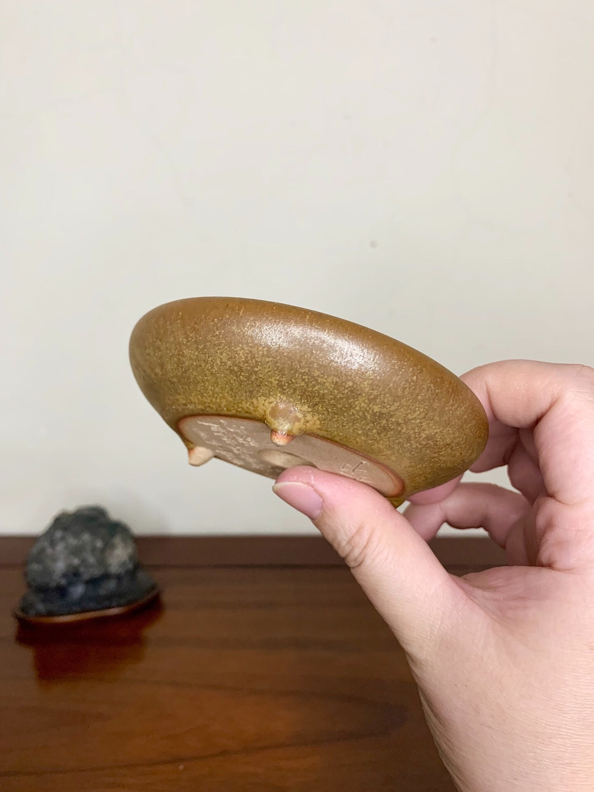 A hand holds a glossy, olive-green glazed pot with three small feet and a smooth, rounded rim.