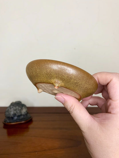 A hand holds a glossy, olive-green glazed pot with three small feet and a smooth, rounded rim.