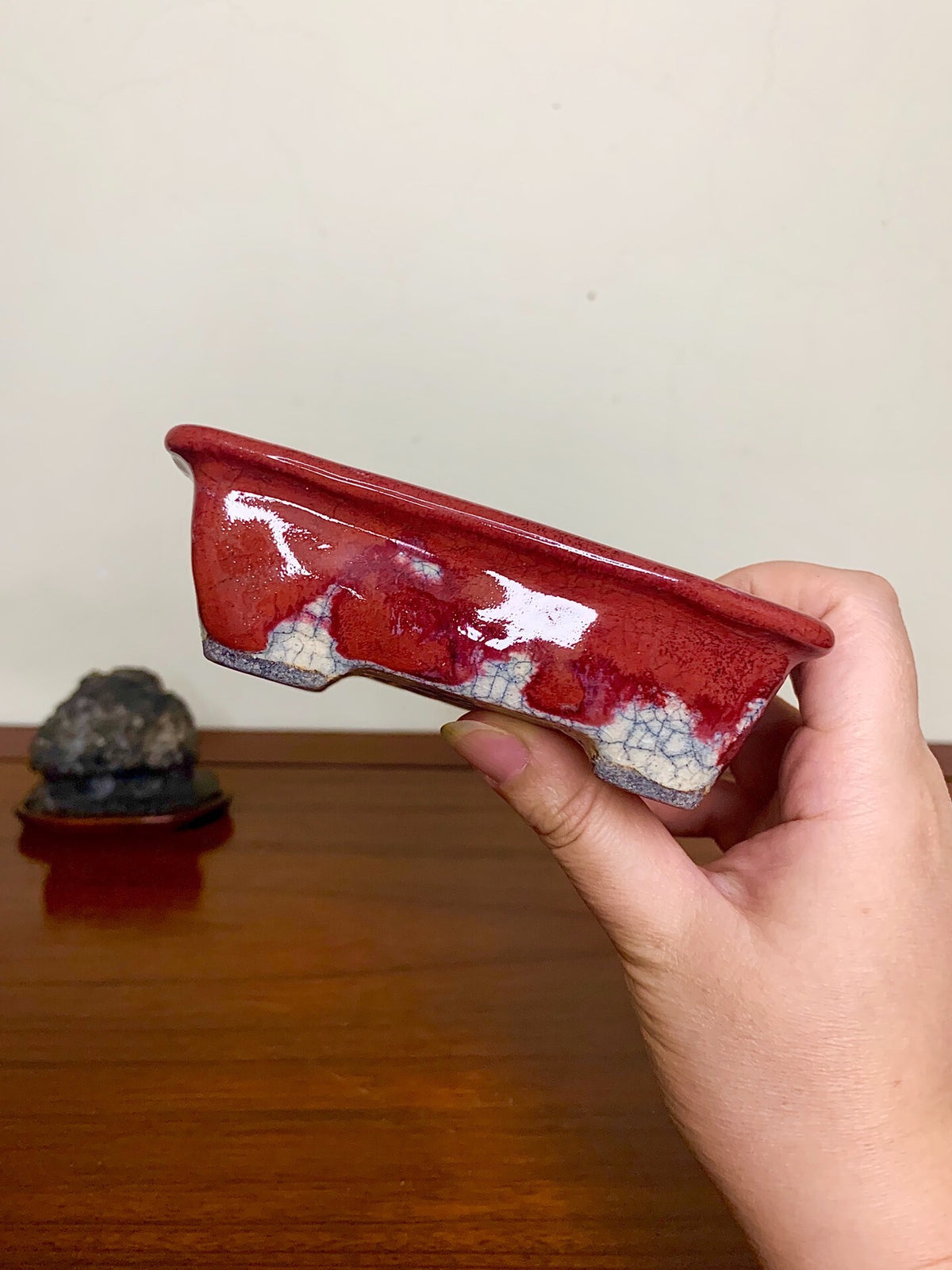A hand holds a glossy red glazed rectangular pot with white crackle pattern on its base.
