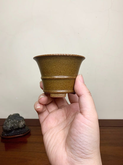 A hand holds a small, olive-green glazed ceramic pot with a flared rim and two horizontal ridges.