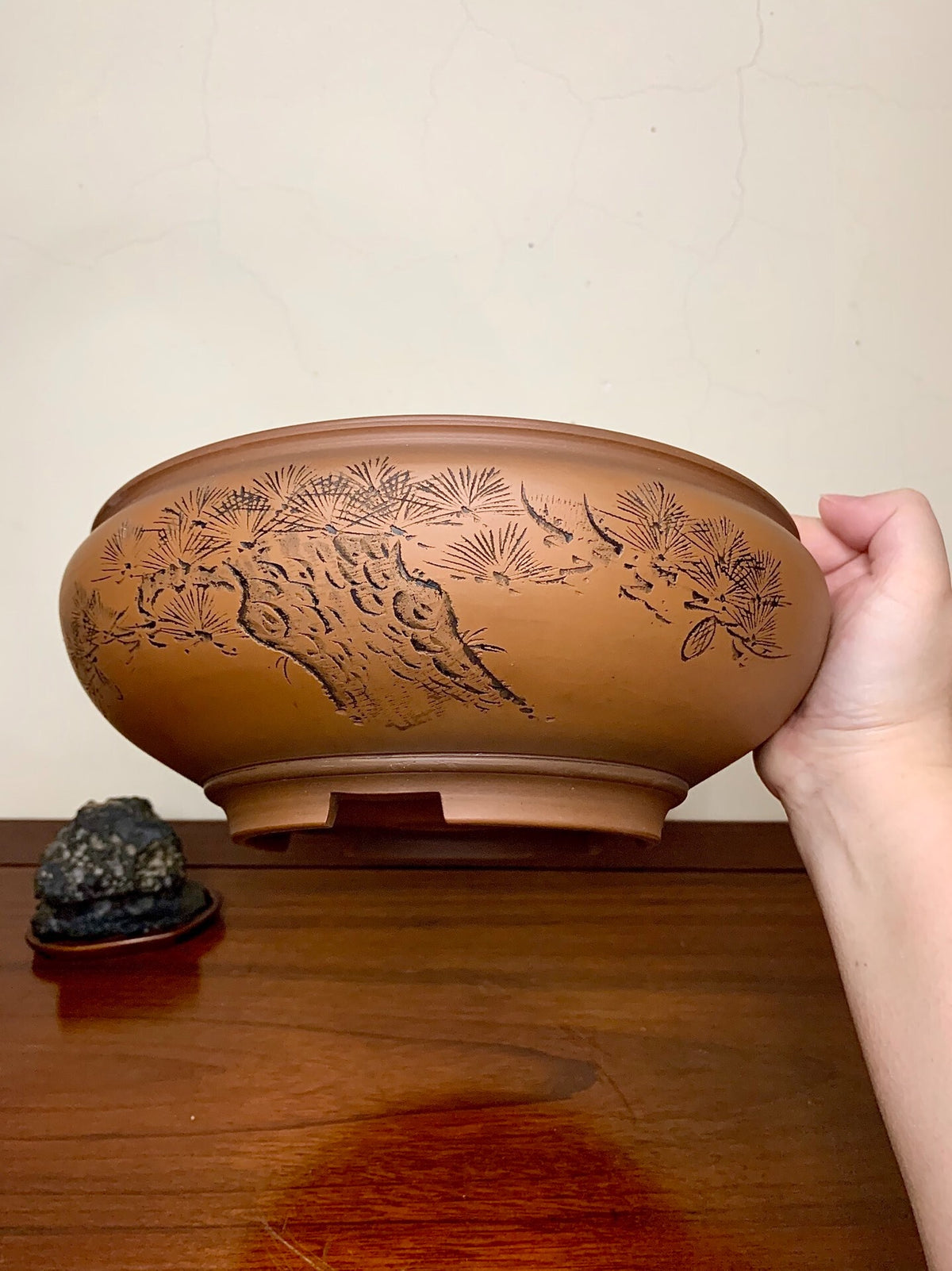 A hand holds a brown glazed pot with carved pine and rock design, beside a dark stone on wood.