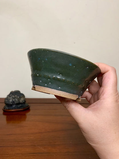 A hand holds a dark green glazed pot with blue speckles and a dotted line near the rim.