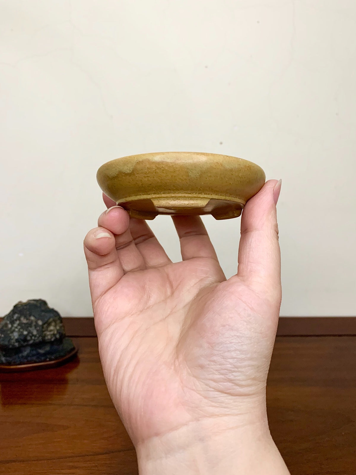 A hand holds a small, shallow, glazed brown ceramic pot with a slightly uneven rim.