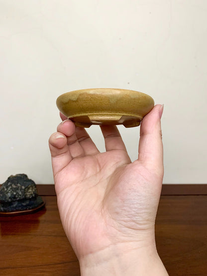 A hand holds a small, shallow, glazed brown ceramic pot with a slightly uneven rim.