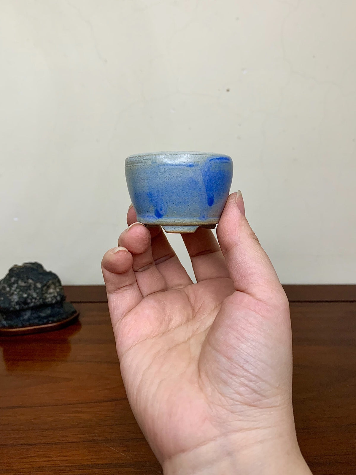 A hand holds a small, blue-glazed ceramic cup with a glossy finish and uneven glaze drips.