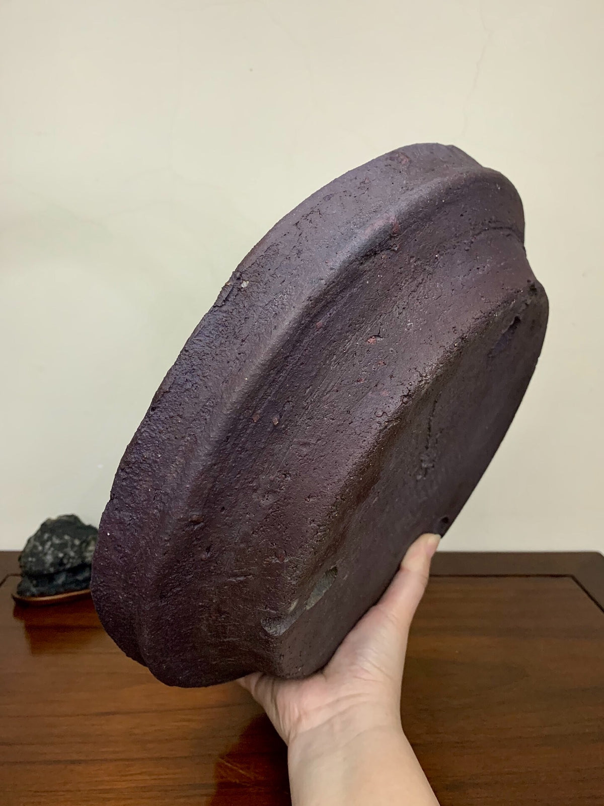 A hand holds a dark, unglazed, roughly textured ceramic pot with a wide, shallow, rounded rim.