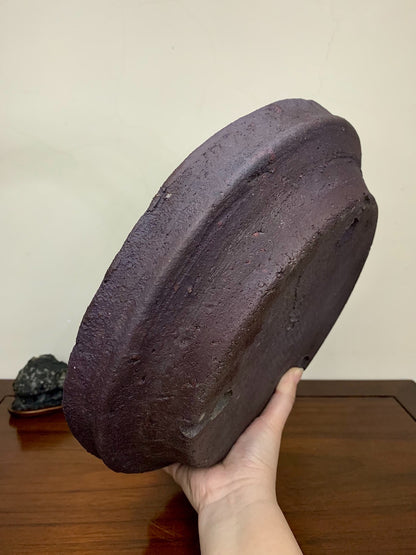 A hand holds a dark, unglazed, roughly textured ceramic pot with a wide, shallow, rounded rim.
