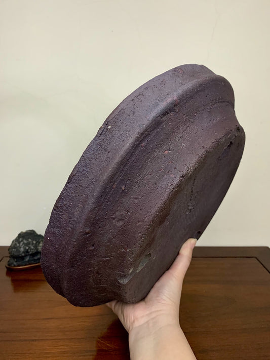 A hand holds a dark, unglazed, roughly textured ceramic pot with a wide, shallow, rounded rim.