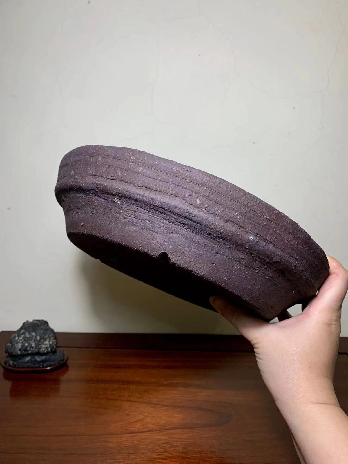 A hand holds a dark brown, unglazed ceramic pot with horizontal ridges and a small hole.