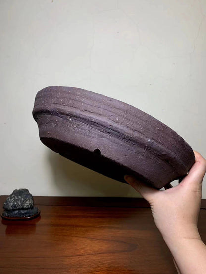 A hand holds a dark brown, unglazed ceramic pot with horizontal ridges and a small hole.