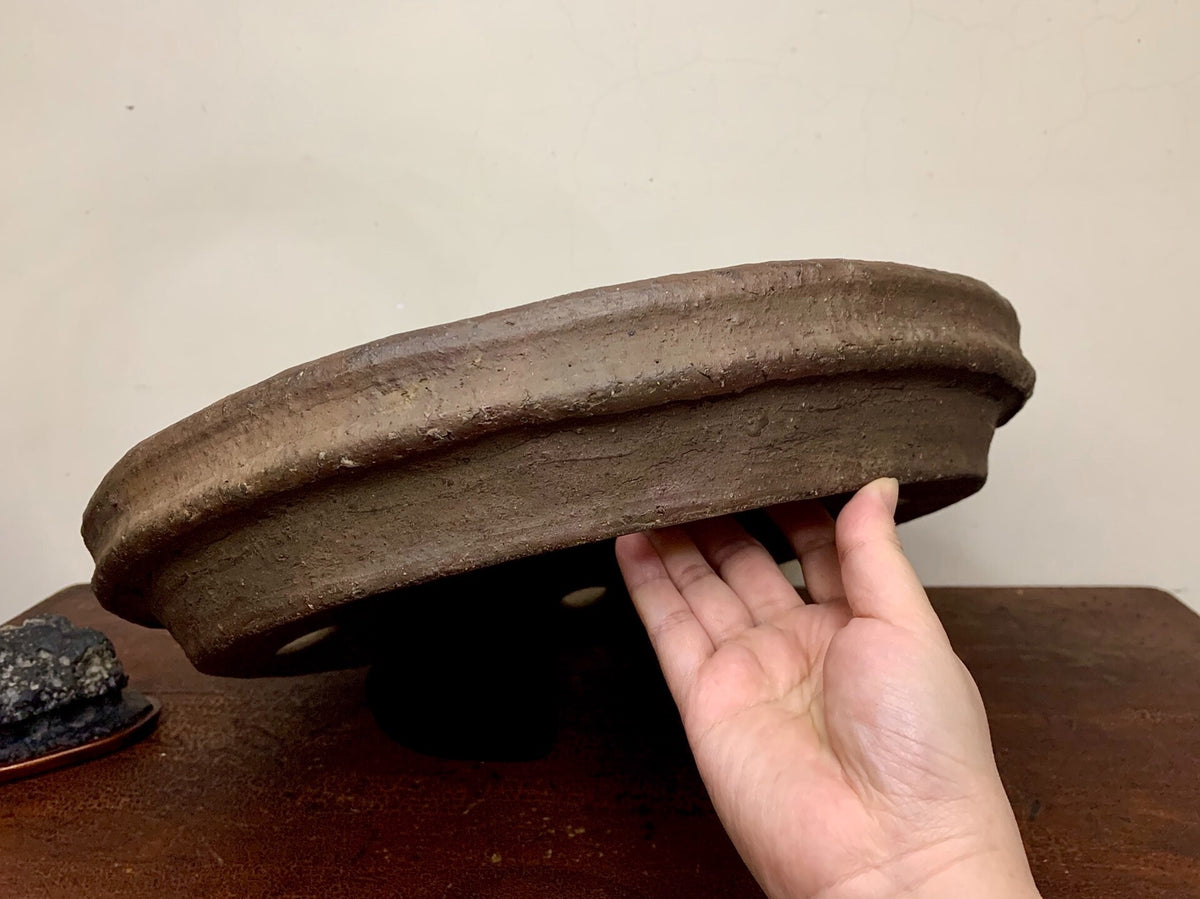 A hand holds a dark brown, unglazed, shallow ceramic pot with a thick rim.