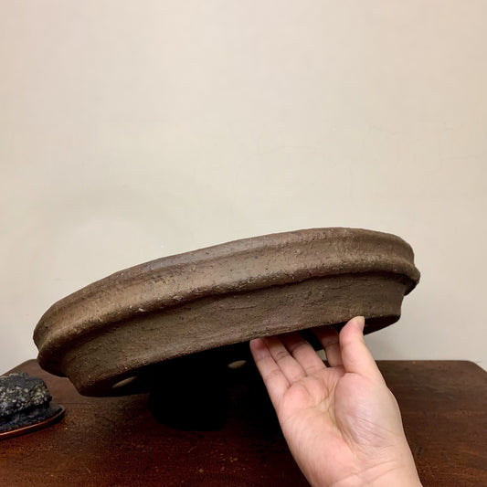 A hand holds a dark brown, unglazed, shallow ceramic pot with a thick rim.