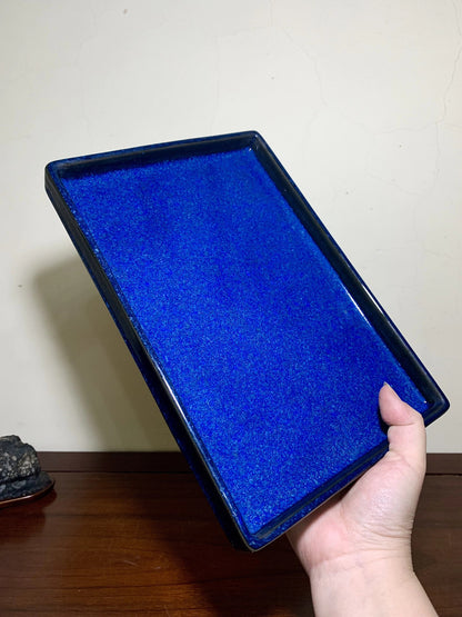 A hand holds a rectangular, glossy cobalt blue glazed tray with a slightly raised rim.