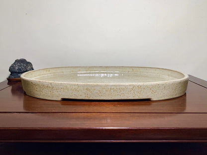 A light beige, oval ceramic tray with speckled glaze sits on a wooden surface.