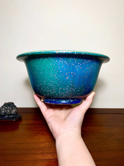 A hand holds a glossy, speckled blue-green ceramic pot with a flared rim.