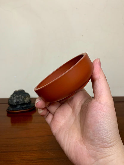 A hand holds a small, round, unglazed terracotta pot with a smooth, matte, reddish-brown surface.