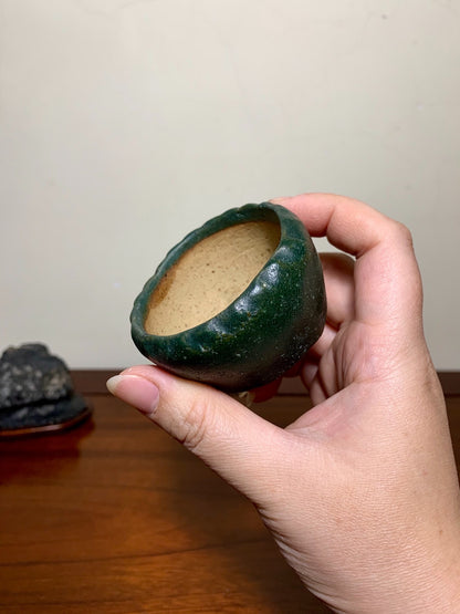 A hand holds a small, dark green glazed pot with a rough, uneven rim and light brown interior.