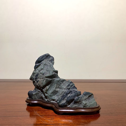 A dark grey, rough-textured rock with layered striations sits on a polished wooden base.