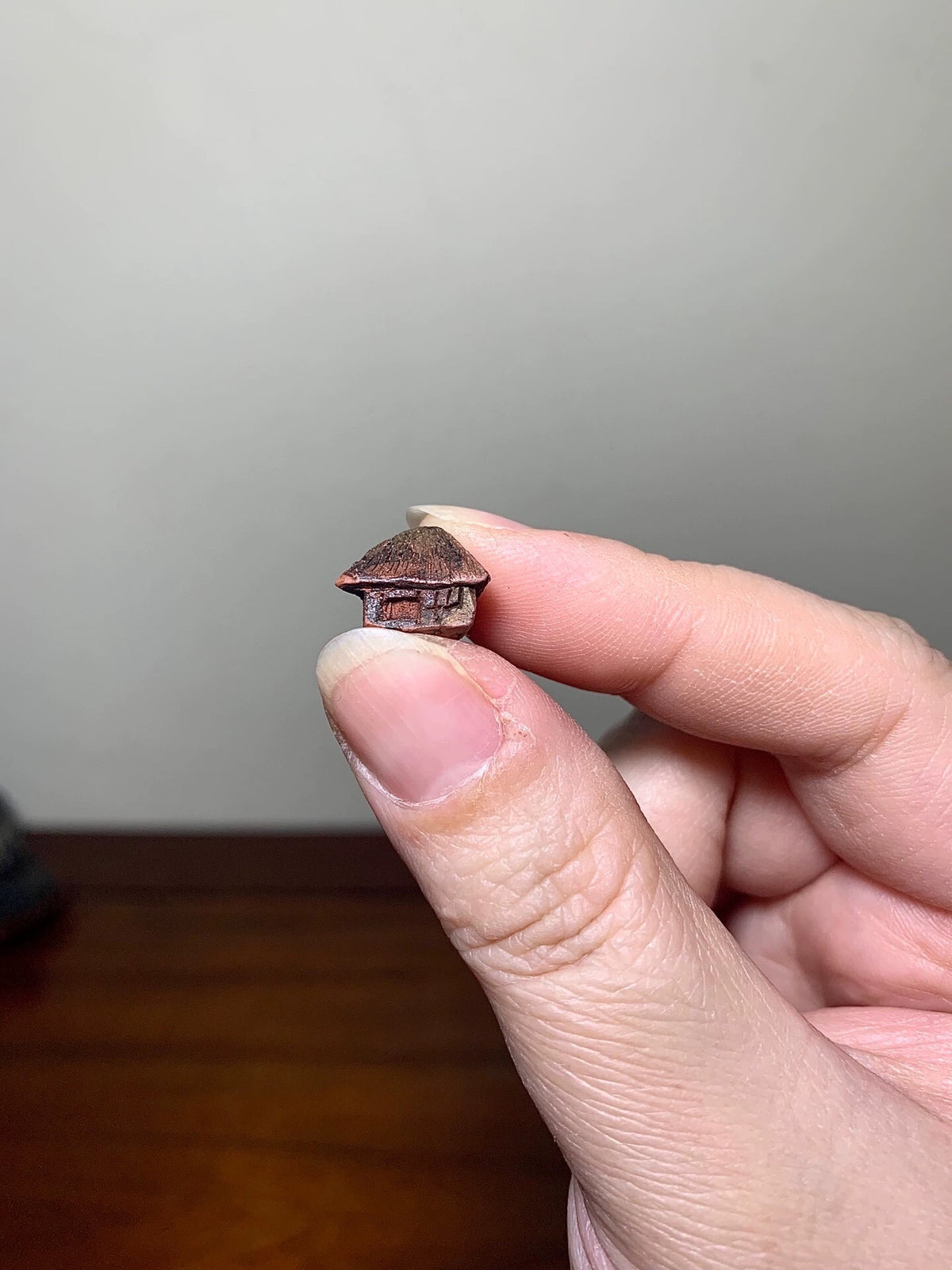 A tiny, dark brown, glazed ceramic house with a thatched roof, held between fingers.