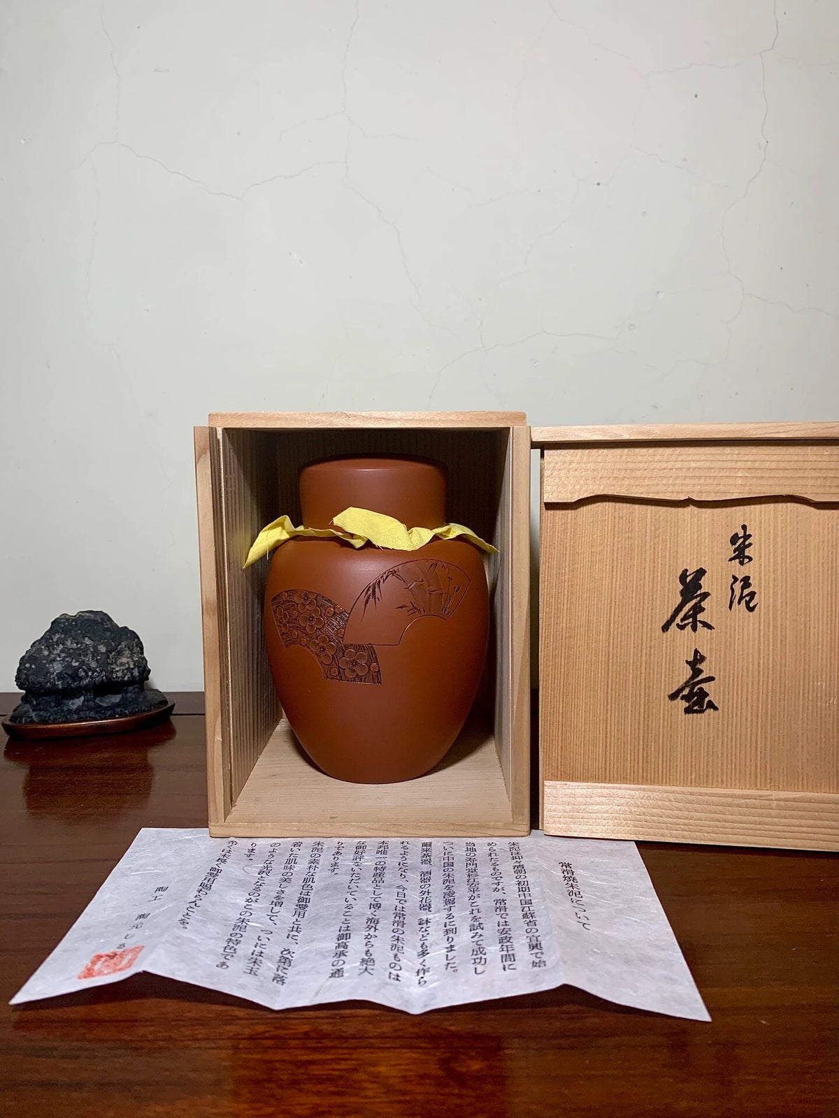 A brown glazed pot with a tree design, wrapped in yellow cloth, sits in a wooden box.