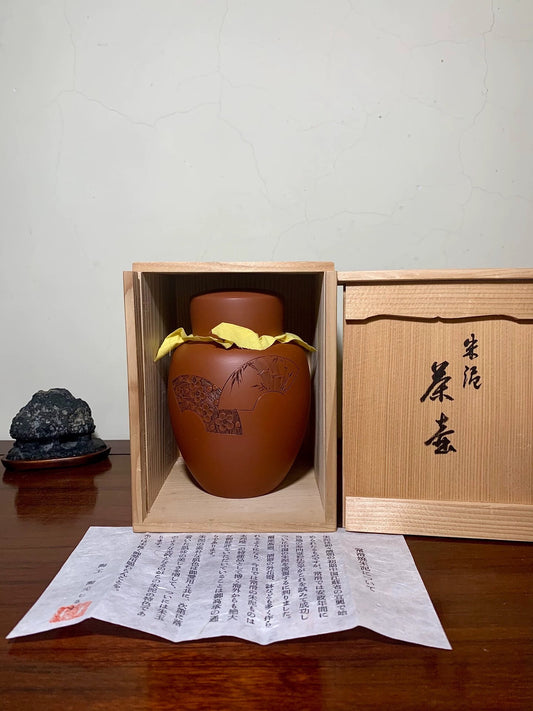 A brown glazed pot with a tree design, wrapped in yellow cloth, sits in a wooden box.