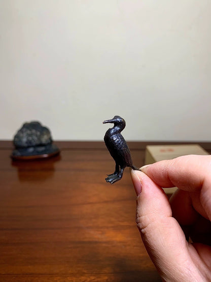 A small, dark, matte-finished bird figurine held between fingers, detailed with textured feathers.