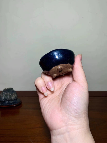 A hand holds a small, glossy dark blue ceramic pot with a brown unglazed base and three small feet.