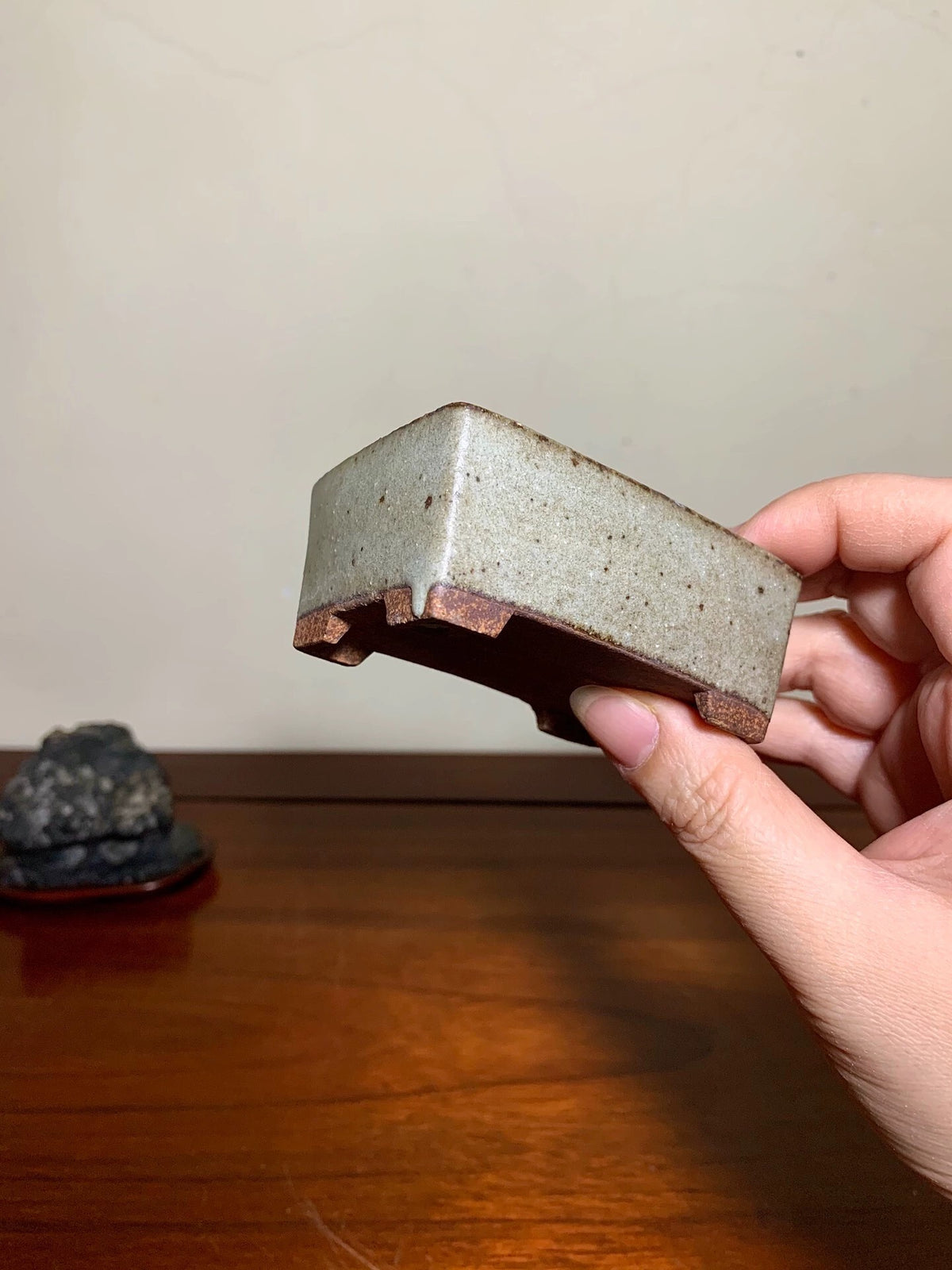 A hand holds a small, rectangular, grey-glazed ceramic pot with unglazed brown rim and base.