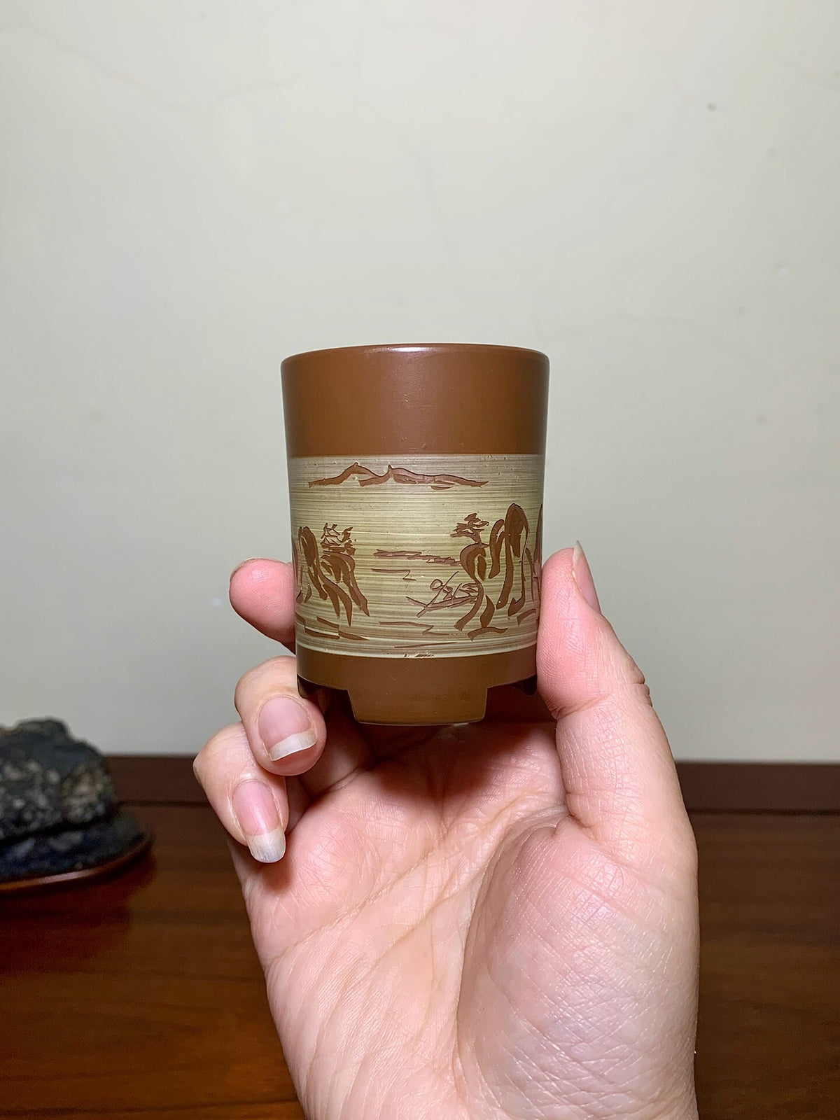 A hand holds a small brown ceramic pot with a beige band showing engraved horses and mountains.