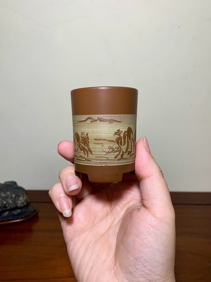 A hand holds a small brown ceramic pot with a beige band showing engraved horses and mountains.