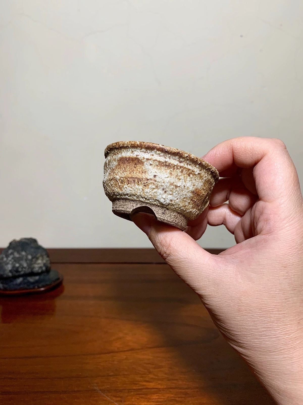 A hand holds a small, rustic, unglazed ceramic cup with brown and beige mottled glaze.