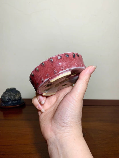 A hand holds a small, deep red glazed pot with metallic studs around its rim.