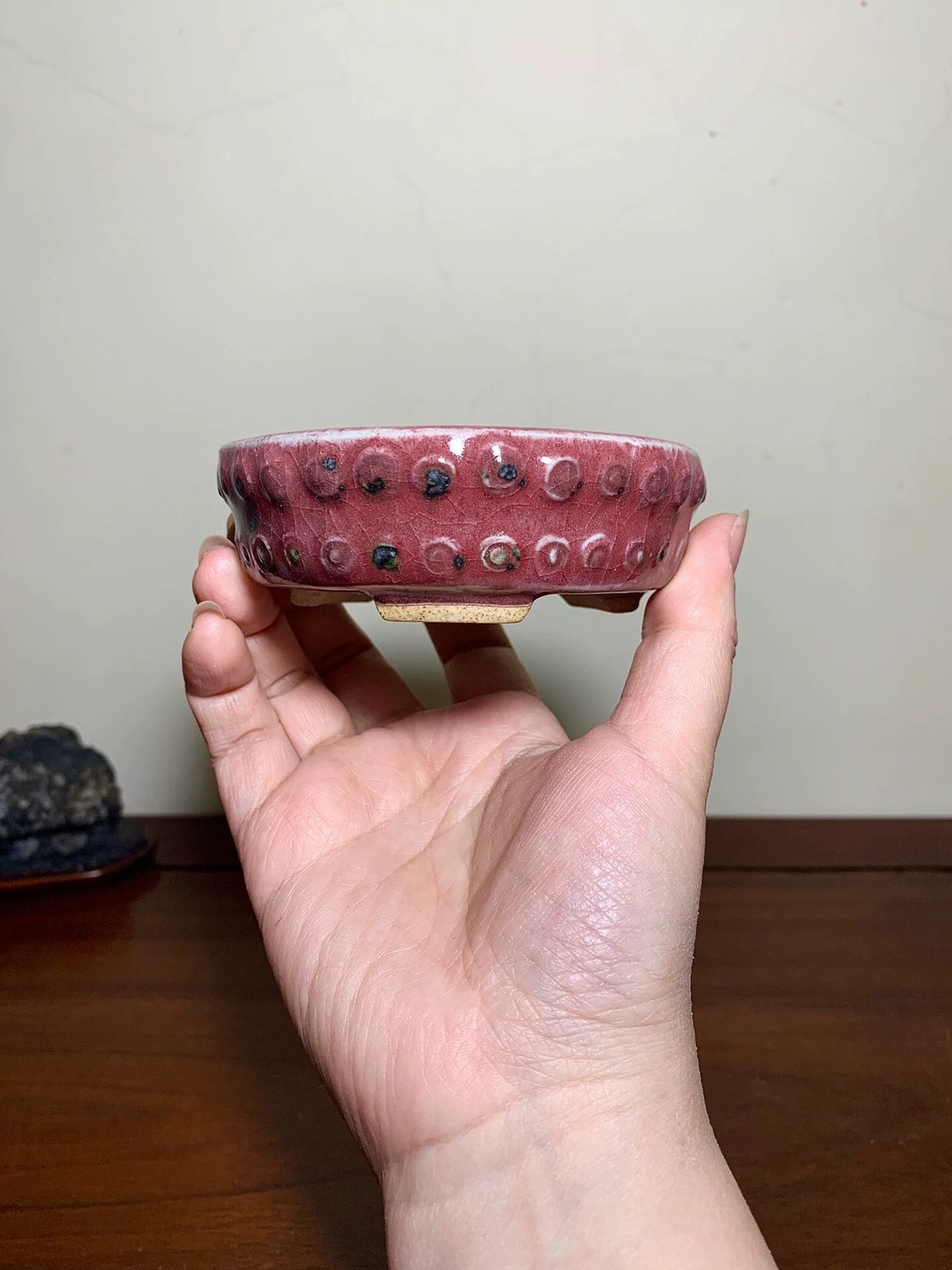 A hand holds a small, deep red glazed pot with raised circular bumps and dark speckles.