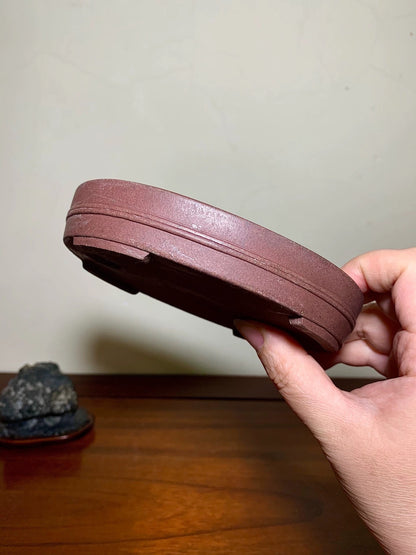 A hand holds a reddish-brown, unglazed ceramic pot base with a textured, matte surface and a slightly raised rim.