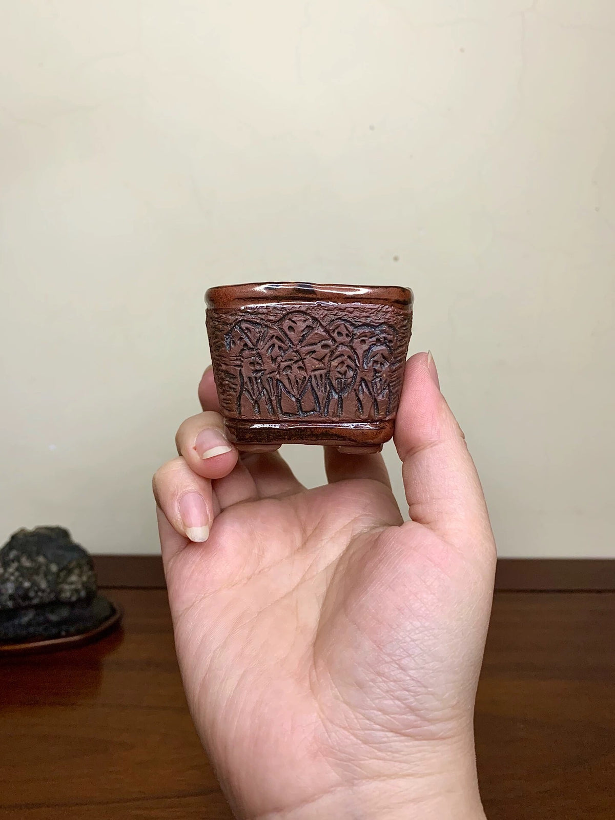 A hand holds a small, dark brown, square pot with intricate carved patterns on its sides.