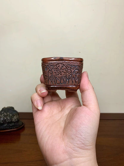 A hand holds a small, dark brown, square pot with intricate carved patterns on its sides.