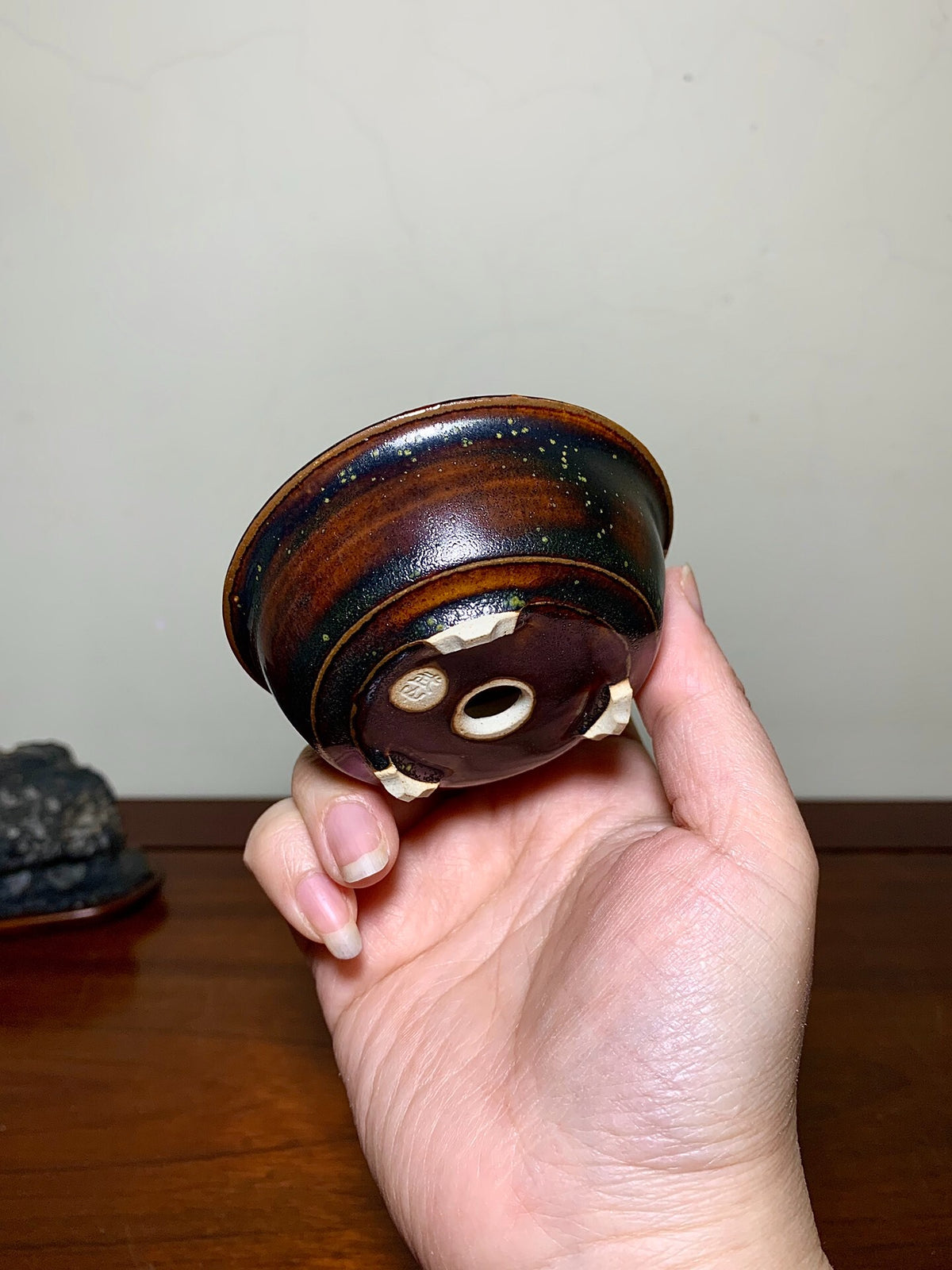 A hand holds a small, glossy, dark brown ceramic pot with a speckled glaze and three white feet.