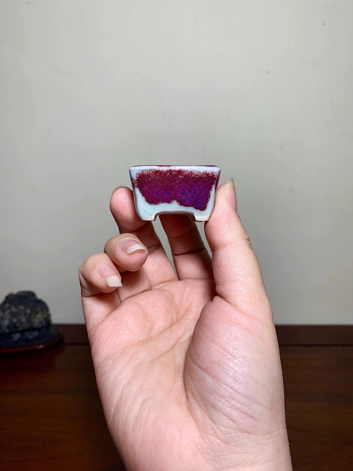 A hand holds a small, square ceramic pot with a glossy white exterior and a vibrant purple and red crystalline interior.