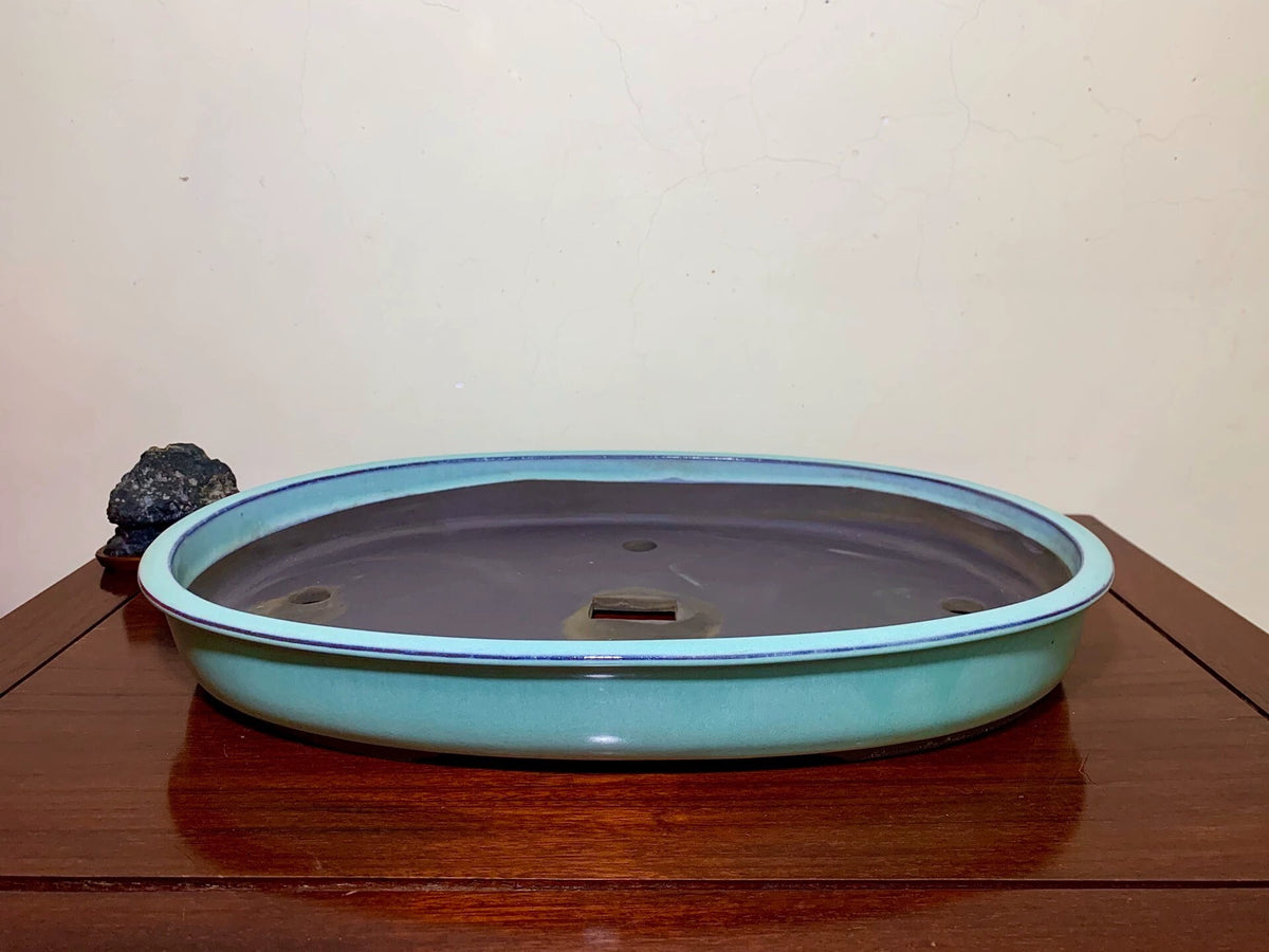 A low, oval glazed pot with a light blue rim and dark grey interior sits on a wooden surface.
